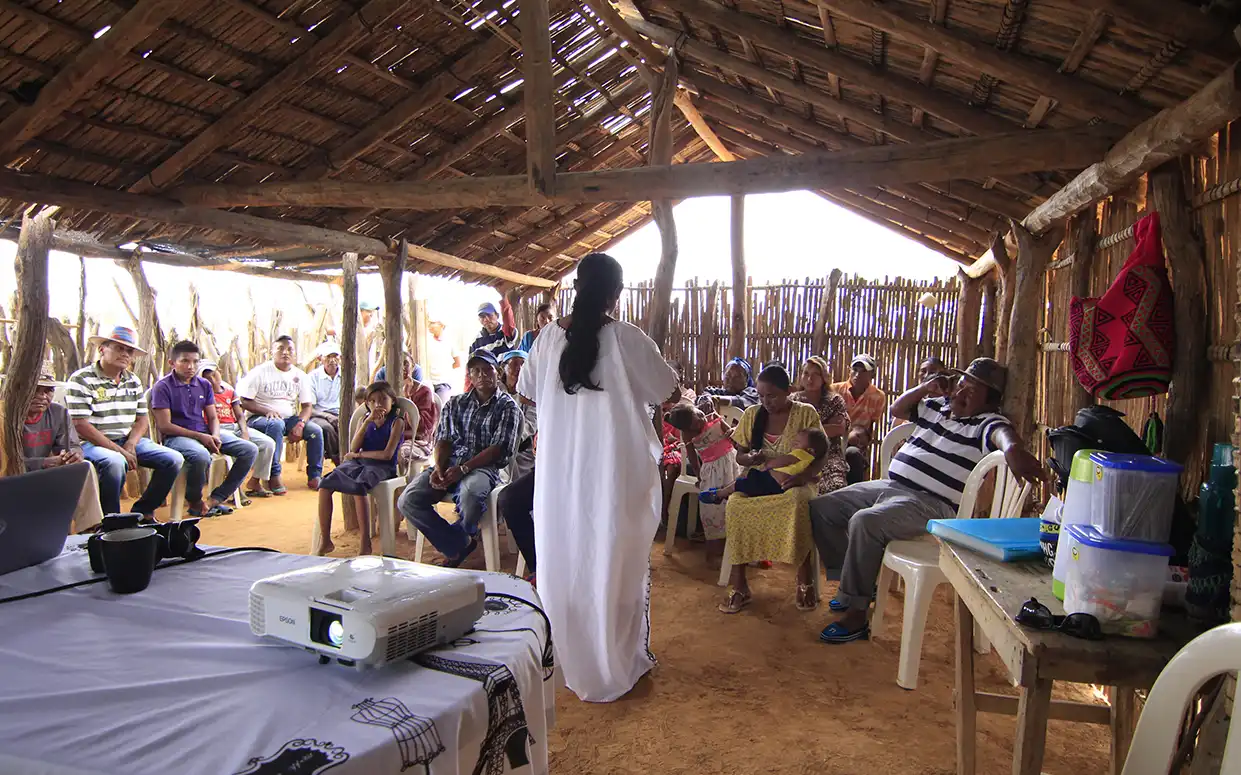 Woman in white presenting in front of locals of a small village in a shack.