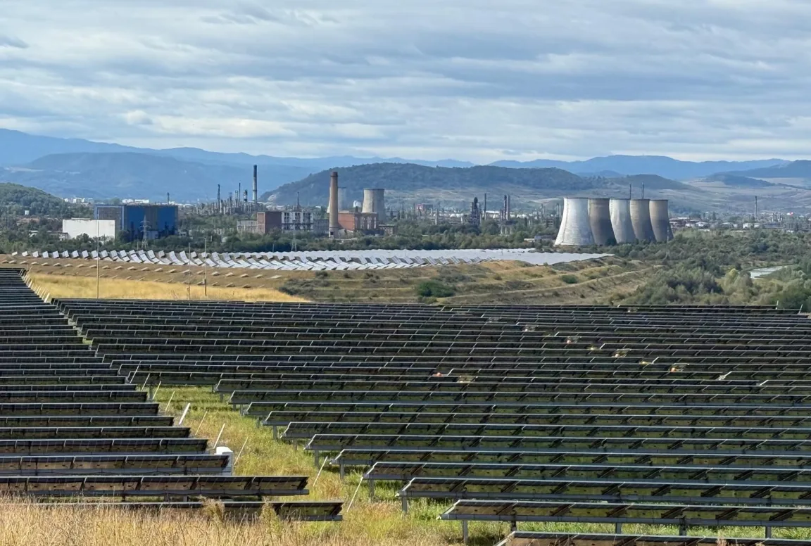 Solar park with rows of photovoltaic panels in front of an industrial site and hills.